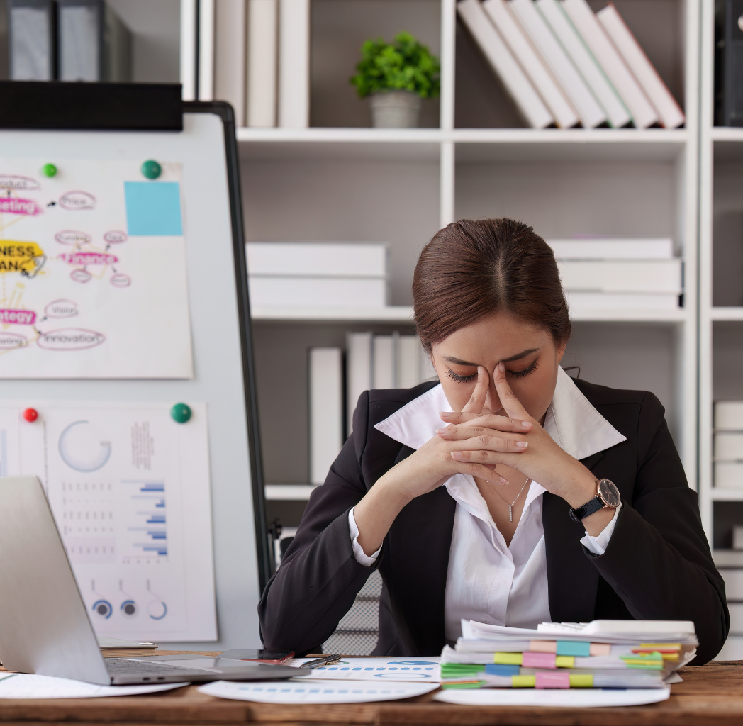 Lady at work looking stressed, sat next to laptop and pile of documents, brdge of nose resting on pointing fingers